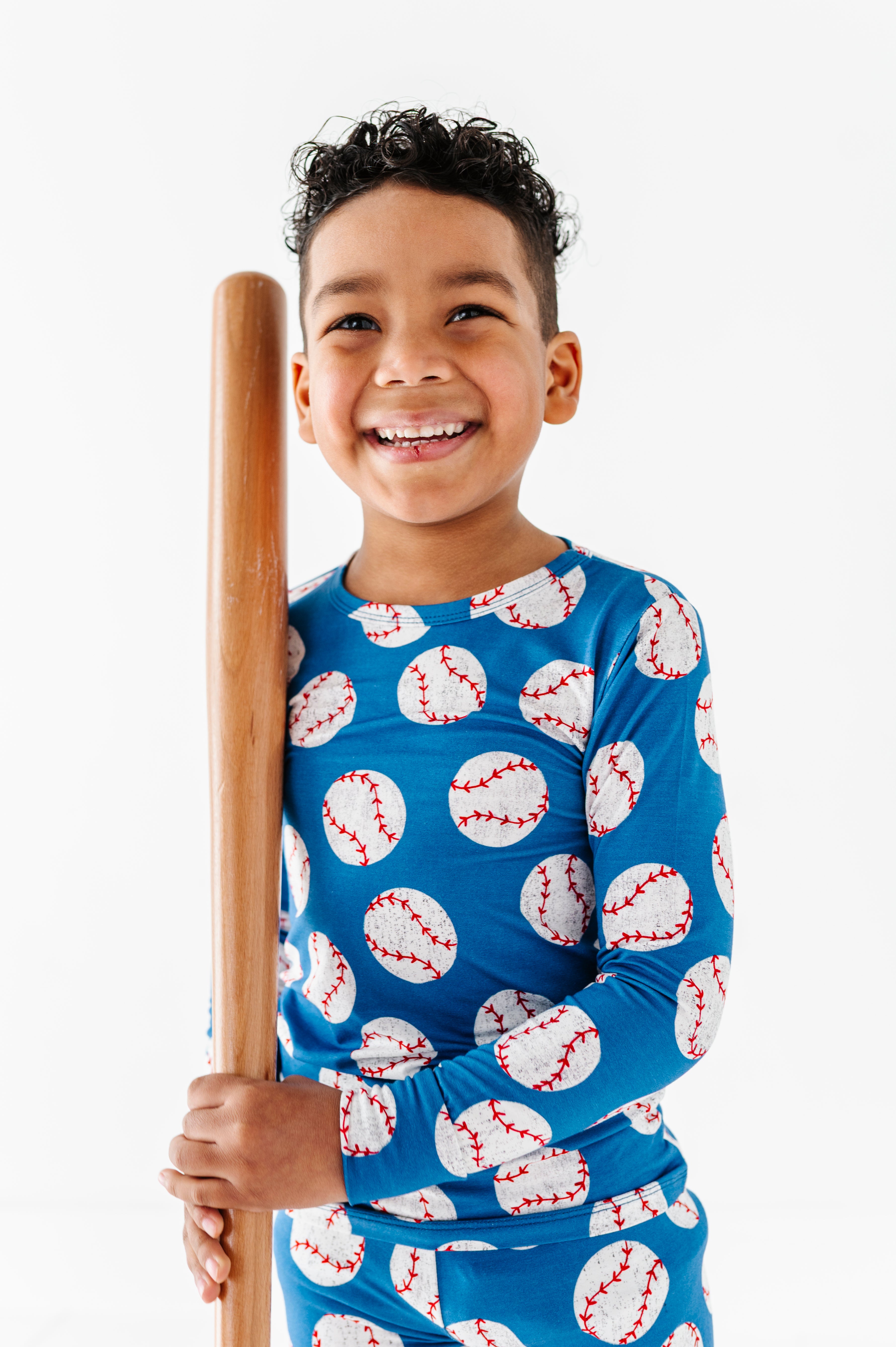 Little boy wearing blue bamboo pajamas featuring a baseball design and holding a wooden baseball bat against a white background
